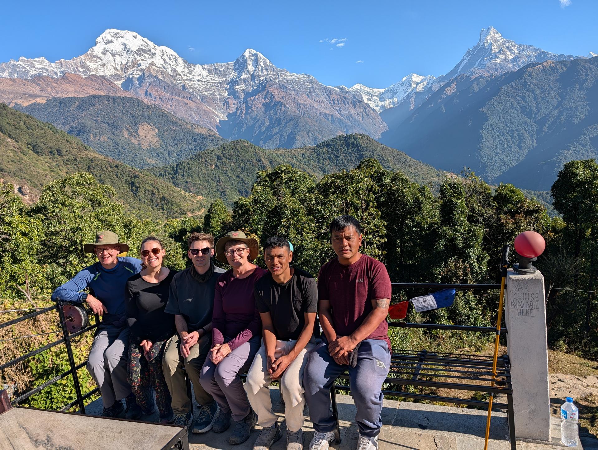 Six people sit together on a terrace railing in front of a sweeping Himalayan panorama of green hills and snowy peaks.