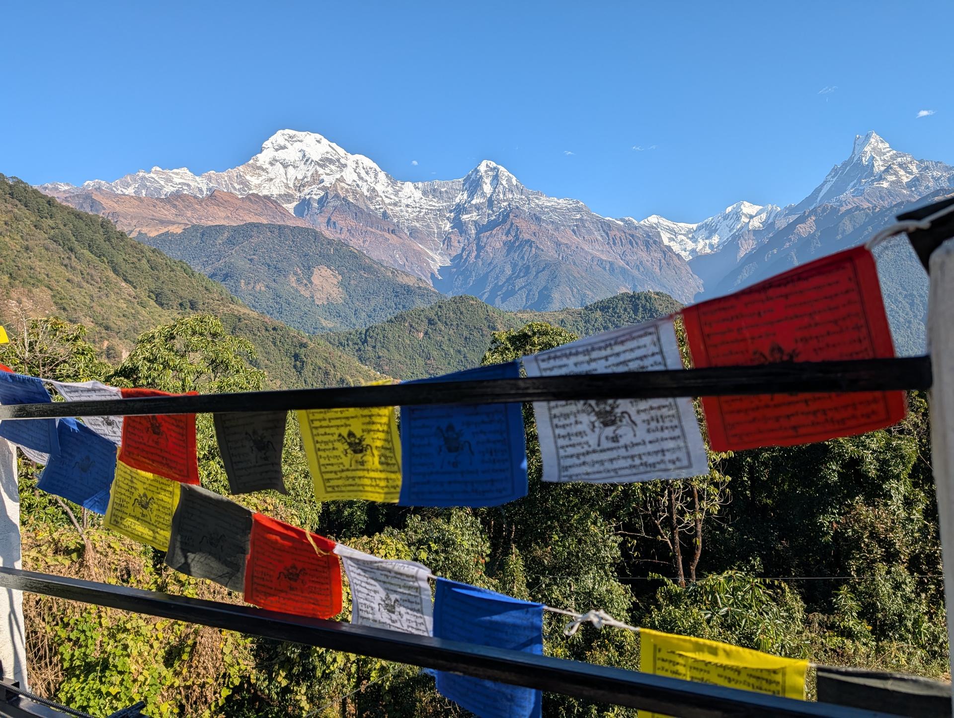 Colorful prayer flags strung across a railing flutter in the foreground, with snow-covered mountains and a deep blue sky beyond.