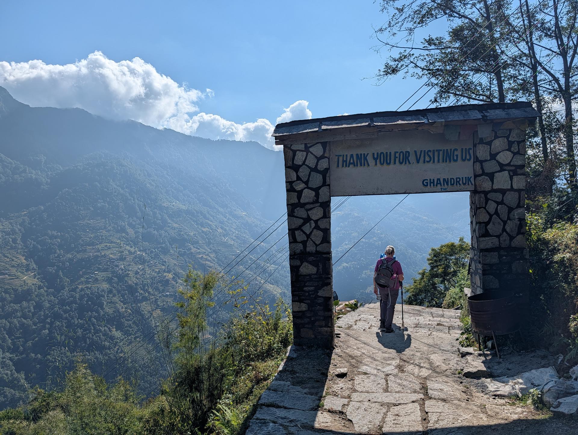 A hiker walks under a stone gateway reading “Thank you for visiting us, Ghandruk,” overlooking a misty mountain valley.