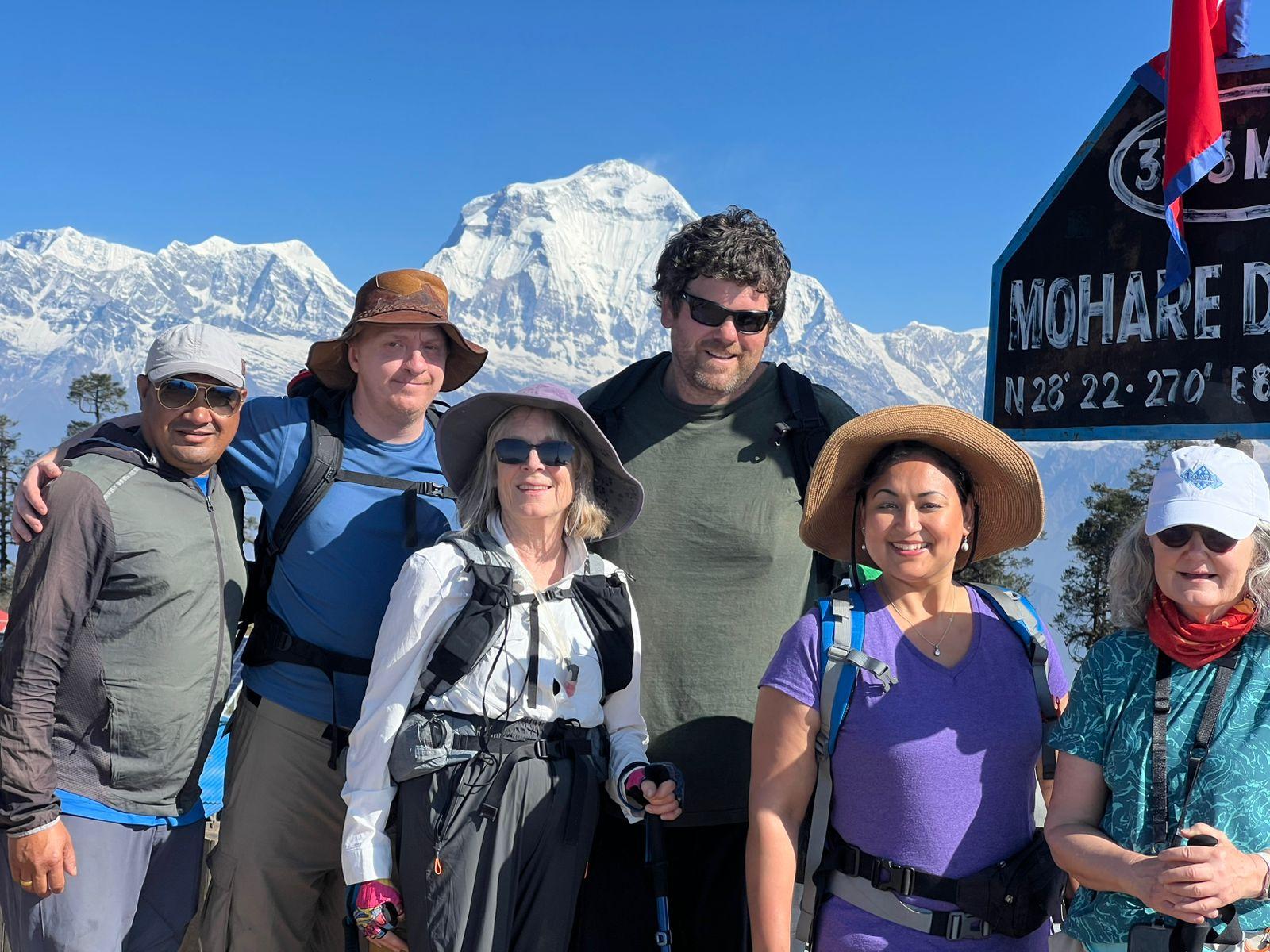 A group of hikers pose beside a sign at a viewpoint, with a massive snow-covered mountain rising behind them.